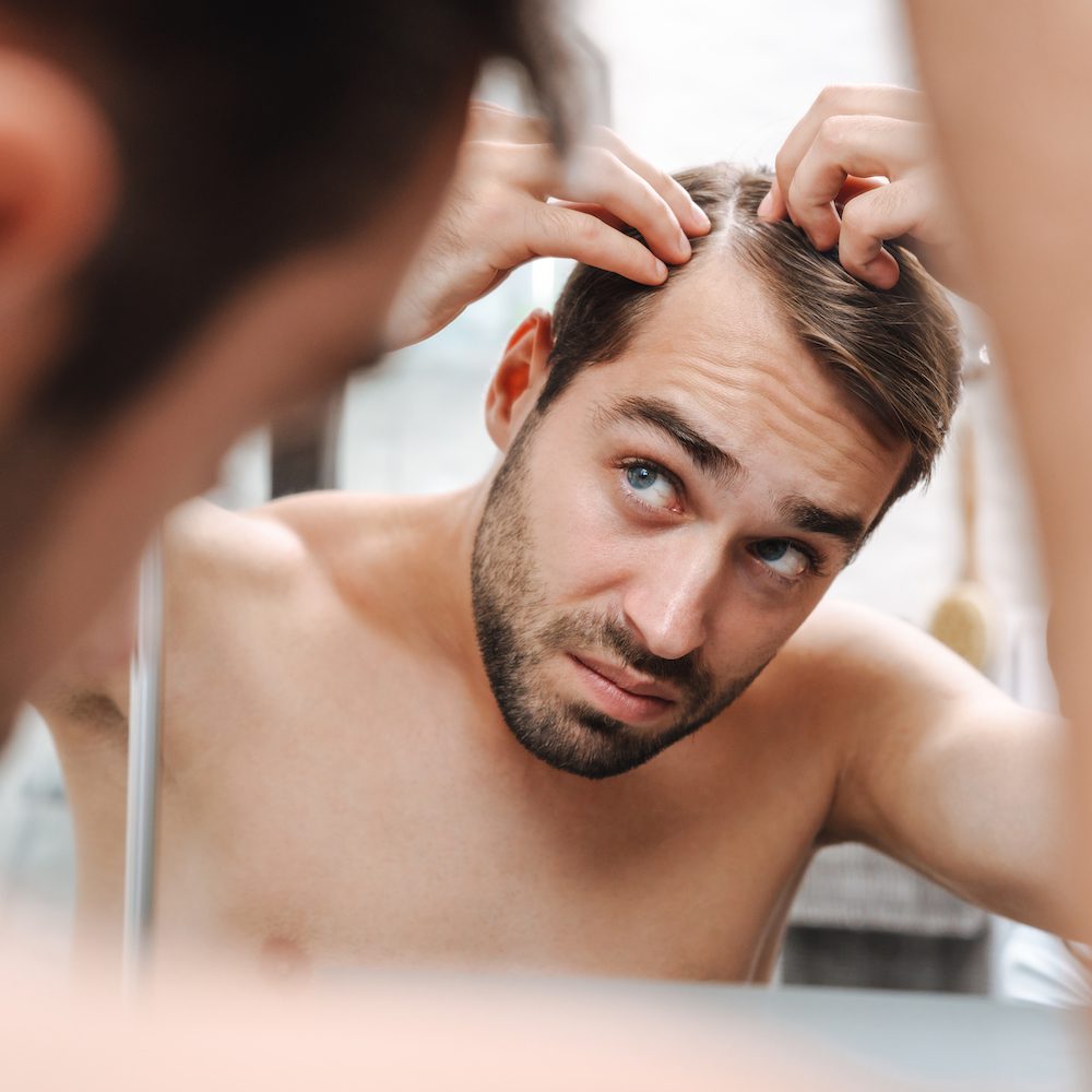 Un homme aux cheveux courts examine son cuir chevelu dans le miroir, l'air inquiet ou pensif. Ses mains effleurent ses cheveux, révélant une légère perte de cheveux sur le dessus – un signe évident de perte de cheveux qu'il constate de plus en plus chaque jour.
