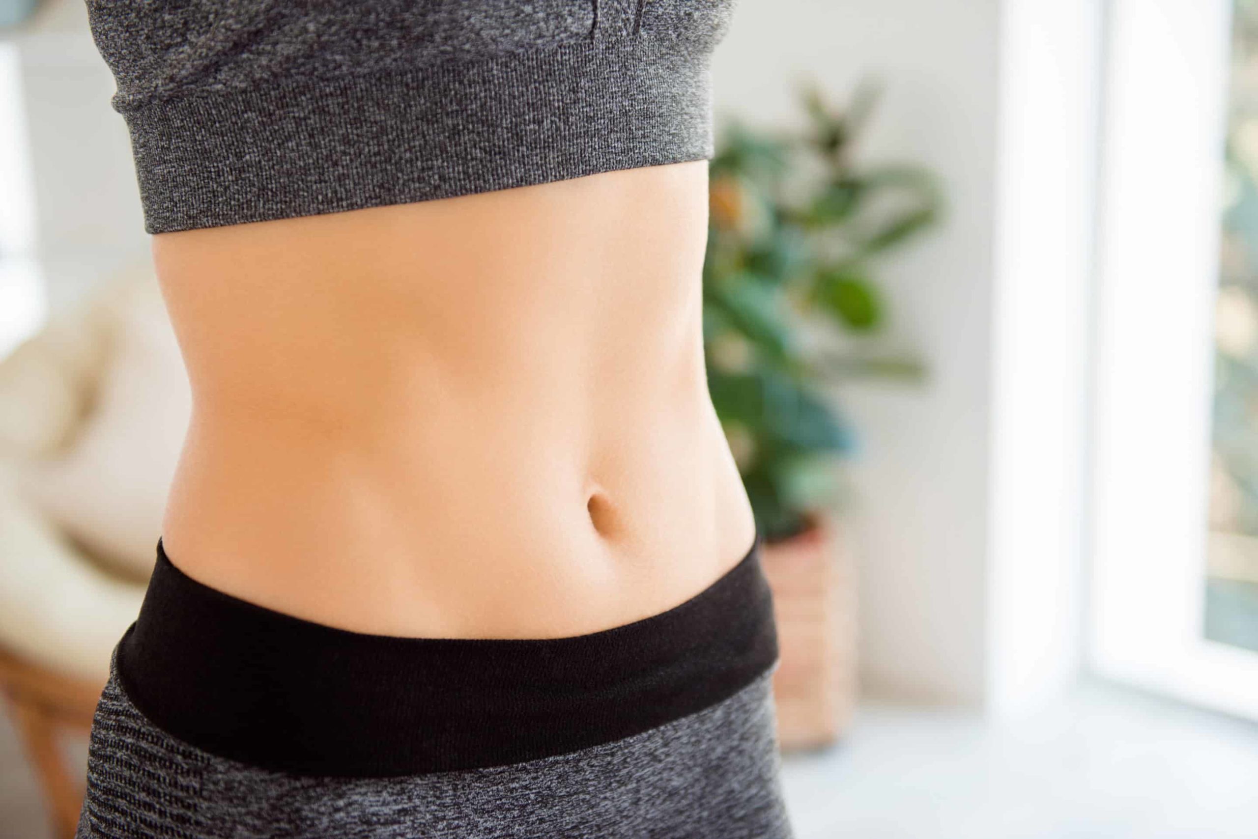 Gros plan sur le ventre d'une personne en tenue de sport grise, mettant en valeur son ventre plat et sa taille ferme. En arrière-plan, une pièce baignée de lumière naturelle et une plante apparaissent légèrement floues.