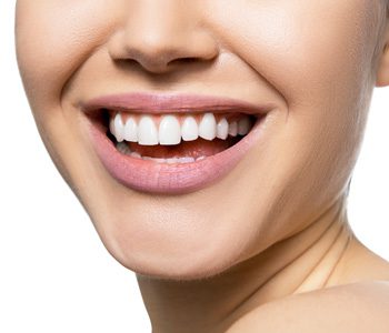 A close-up of a person smiling, showcasing the results of cosmetic dentistry. The lips are slightly parted, and the skin is smooth and well-lit. The image focuses on the lower half of the face, capturing a cheerful expression with perfectly aligned teeth.