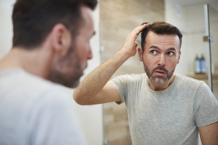Un homme aux cheveux noirs courts et à la barbe, vêtu d'un t-shirt gris, examine son reflet dans le miroir de la salle de bain, songeant à des greffes de cheveux tout en se touchant les cheveux. L'arrière-plan révèle des carreaux flous et des articles de toilette assortis.