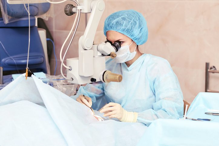 A surgeon in a blue surgical gown, mask, and cap performs a LASIK procedure using a microscope in the operating room. The space is equipped with various medical tools and sterile blue sheets carefully covering the patient.