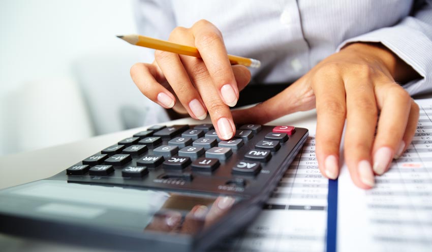 Gros plan d'une personne utilisant une calculatrice d'une main et tenant un crayon de l'autre. L'arrière-plan montre des documents imprimés indiquant une tâche financière ou budgétaire.