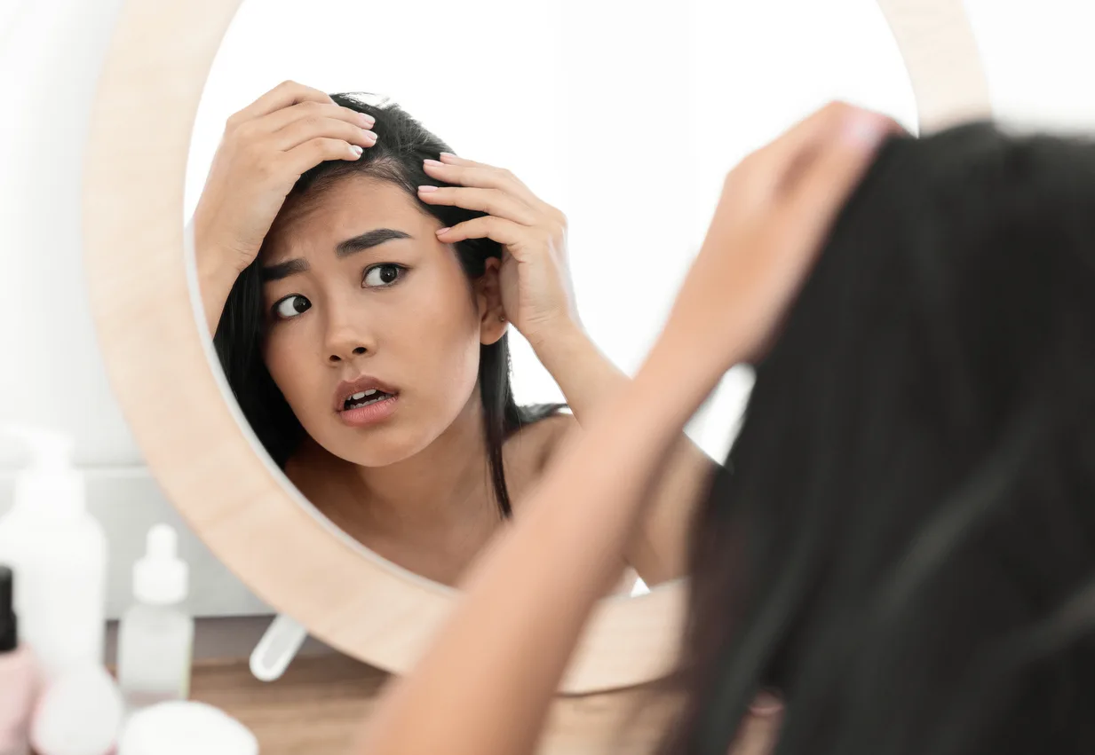 Une femme aux longs cheveux noirs regarde avec inquiétude son reflet dans un miroir rond, songeant à un traitement contre la chute des cheveux. Elle examine son cuir chevelu, touchant ses cheveux des deux mains. Des flacons de produits de soin sont posés sur la table devant elle.