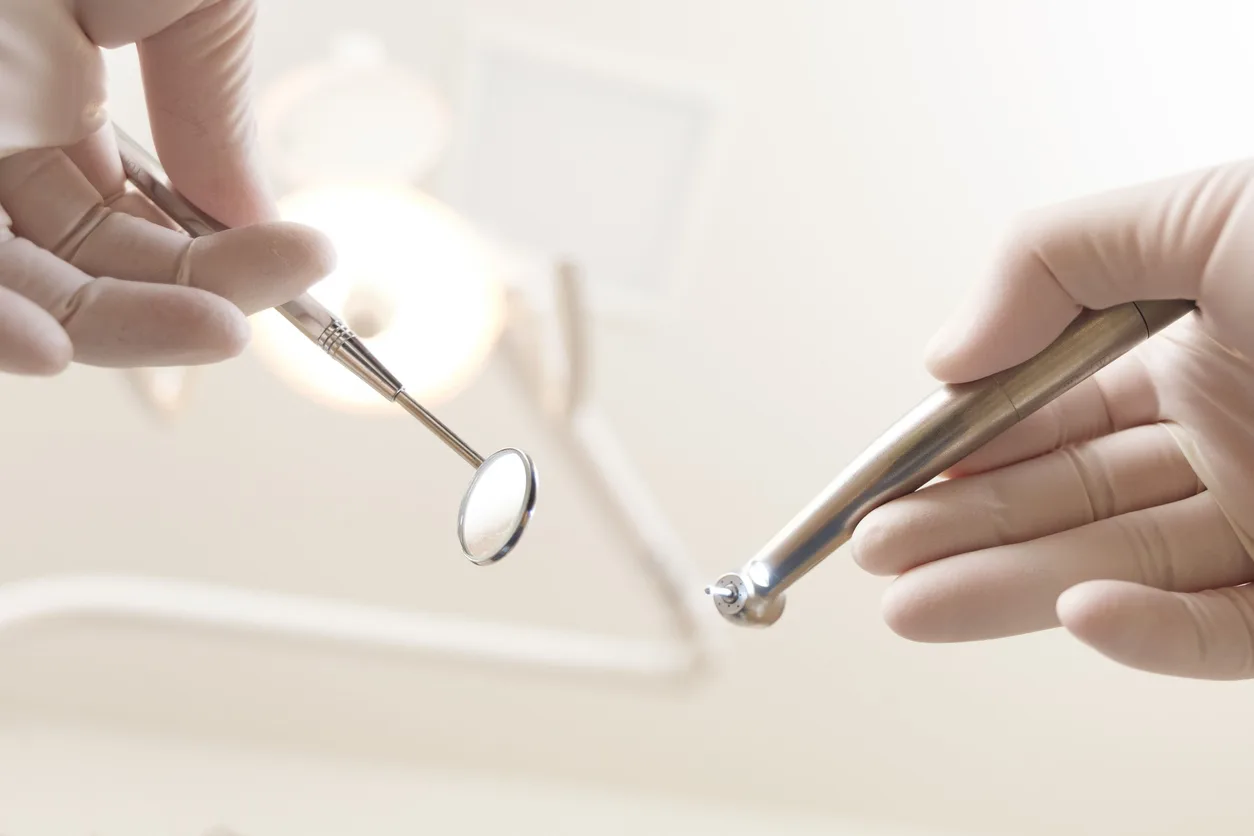 Close-up of gloved hands holding dental tools, including a mirror and drill, against a blurred background of a dental light. The scene suggests a dentist preparing for dental surgery.