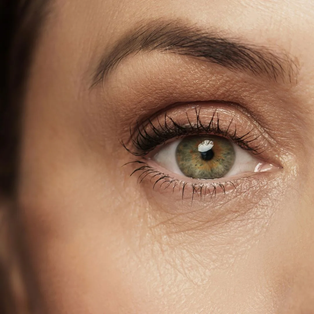 Gros plan sur l'œil d'une personne à l'iris brun clair et aux cils proéminents. La peau autour de l'œil présente une texture et des nuances naturelles, sublimées par un maquillage subtil. Des reflets lumineux sont visibles dans l'œil, laissant entrevoir l'éclat post-chirurgie.