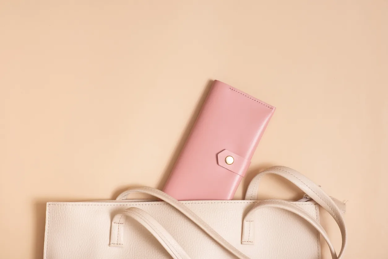A pink wallet with a button closure peeks stylishly from a beige handbag, set against a matching background. The handbag, with its two visible handles, mirrors the precision and elegance often associated with cosmetic surgery, creating a minimalistic and chic composition.