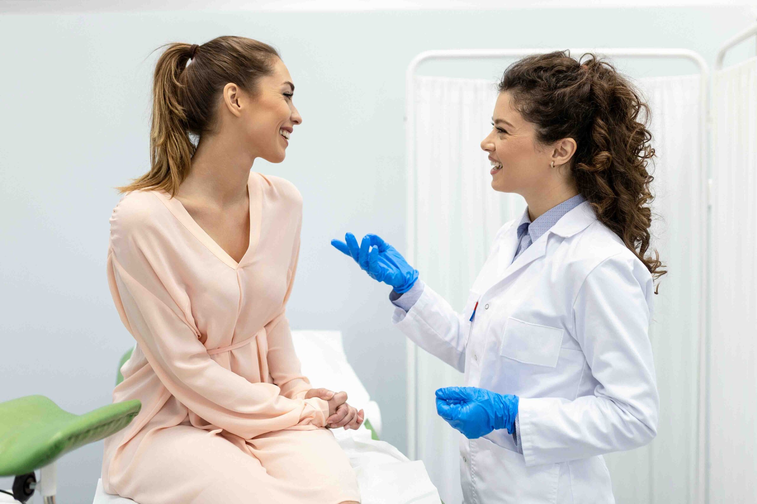 Une femme en robe rose est assise sur une table d'examen médical et sourit à un professionnel de santé portant une blouse blanche et des gants bleus. Elles discutent, un rideau et du matériel médical étant visibles en arrière-plan.