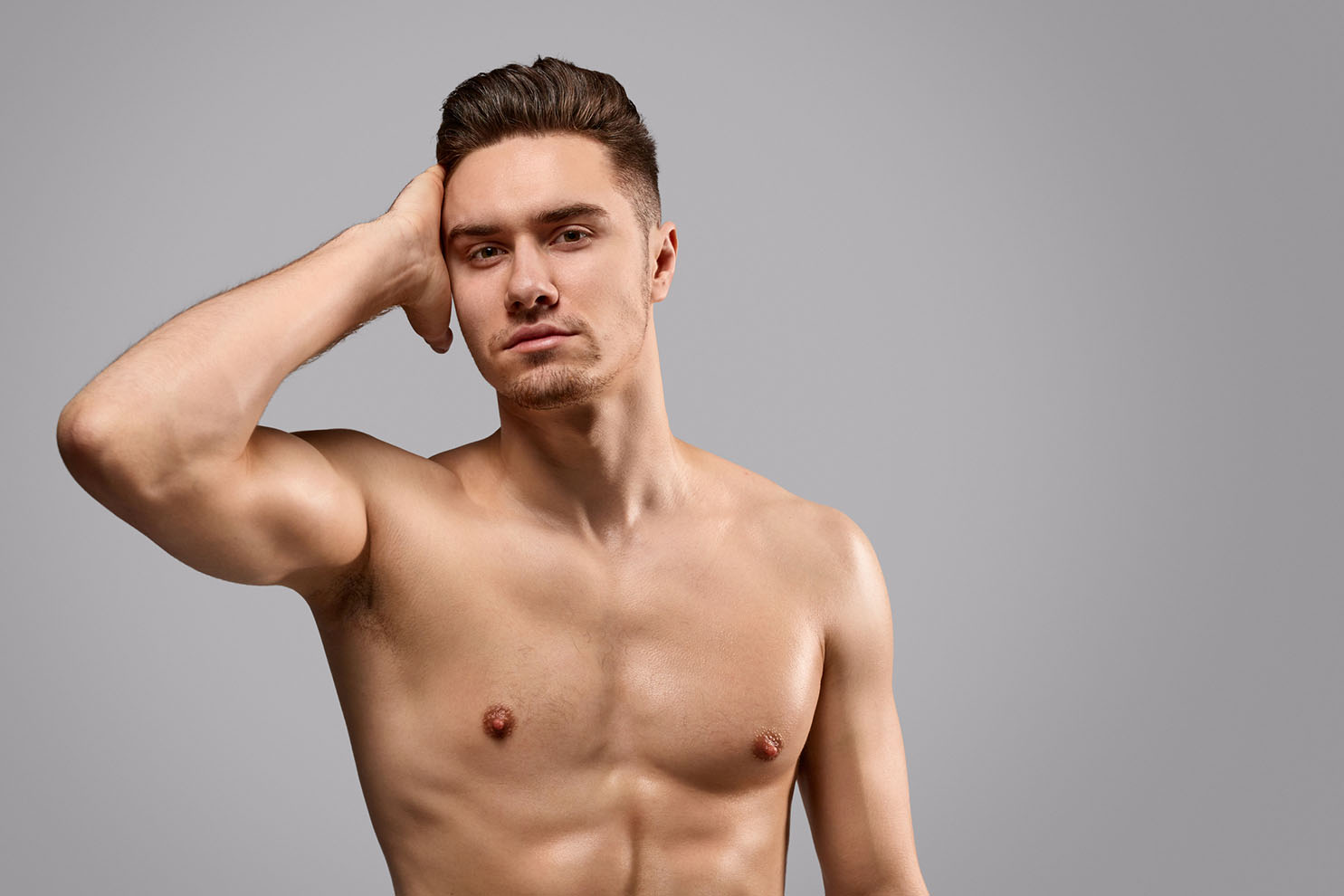 A shirtless man poses against a plain gray background. He has short brown hair and is looking directly at the camera with his right hand touching his head. His expression is neutral.