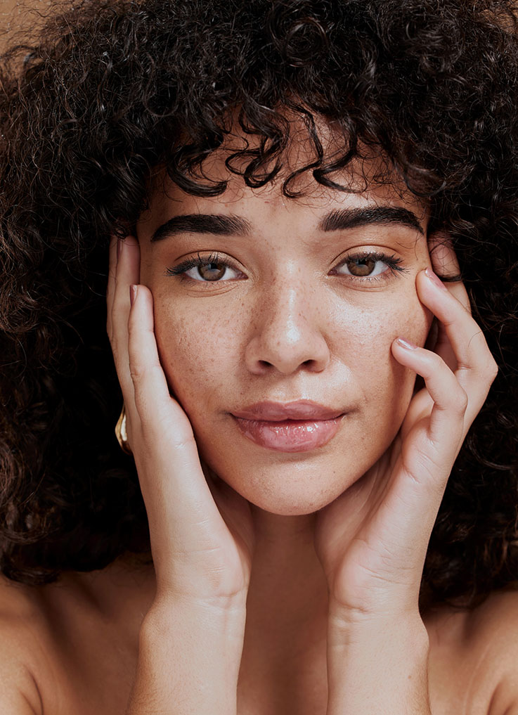 A person with curly hair and freckles holds their face with both hands. They have a calm expression and are looking directly at the camera. The background is neutral, highlighting their facial features.