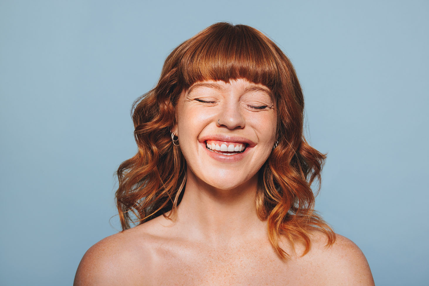 A person with wavy red hair and bangs, eyes closed, smiling widely against a light blue background. They have a freckled complexion and are wearing small hoop earrings.