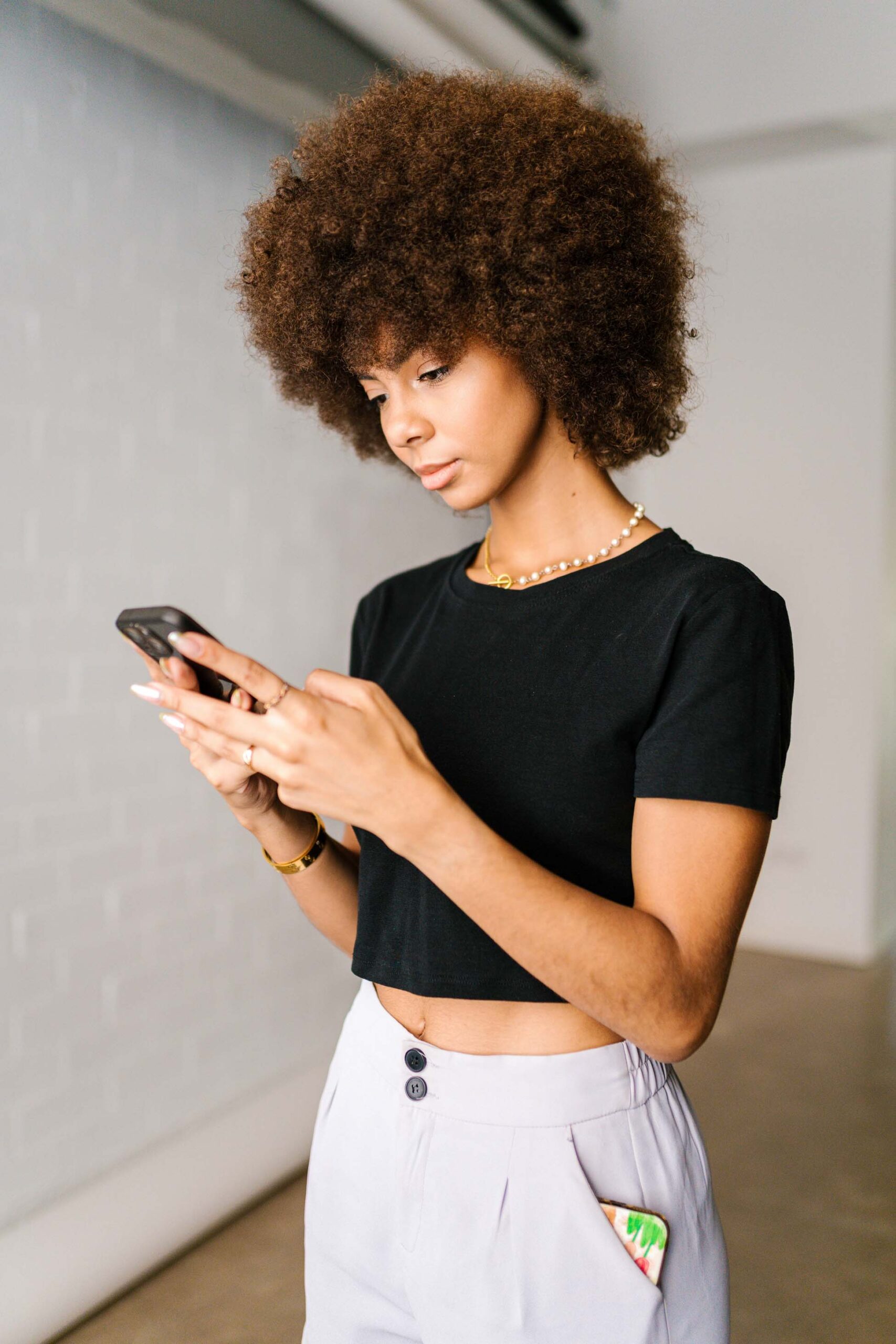 Une femme aux cheveux bouclés se tient à l'intérieur, concentrée sur son smartphone. Elle porte un haut court noir, un pantalon clair et un collier. L'arrière-plan est simple, avec une vue partielle sur un mur.