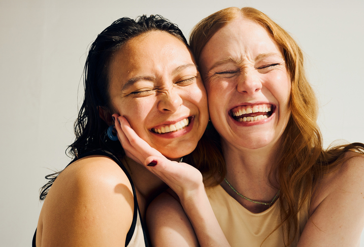 Two women laughing cheek to cheek in bright light, showing big smiles—image for a guide on invislaign costs.